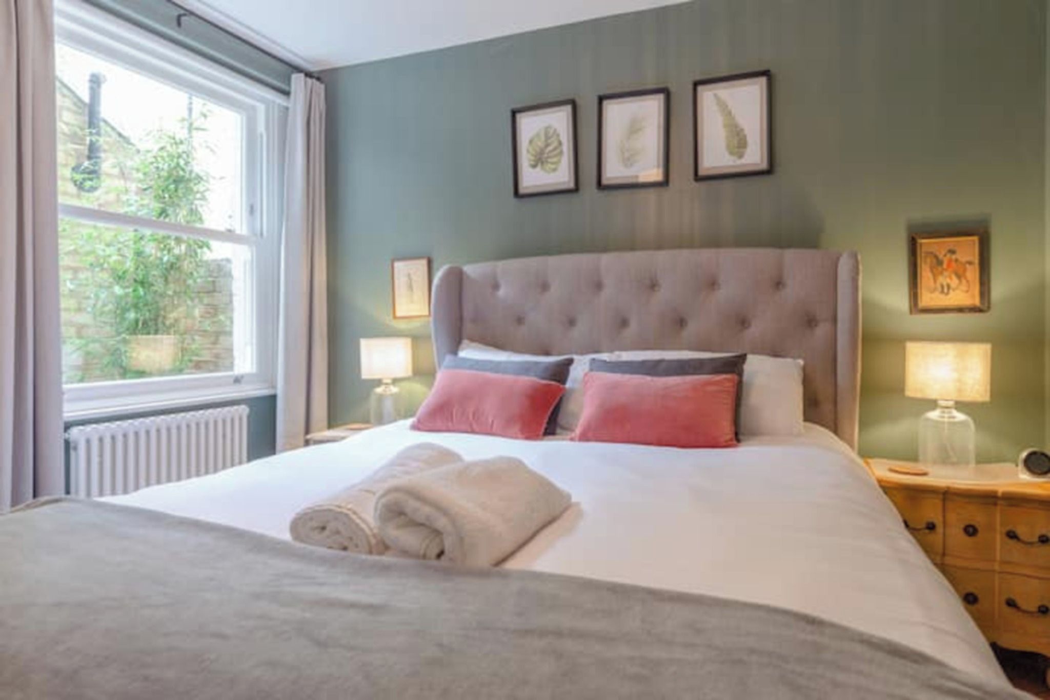 A neatly made bed with a grey duvet and rolled towels, a tufted headboard, and bedside lamps on wooden nightstands.