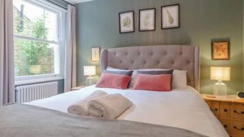 A neatly made bed with a grey duvet and rolled towels, a tufted headboard, and bedside lamps on wooden nightstands.