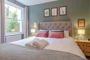 A neatly made bed with a grey duvet and rolled towels, a tufted headboard, and bedside lamps on wooden nightstands. - Cosy stylish 1 bed flat in Fulham w patio (London)