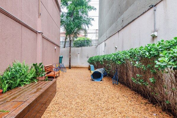 Un espacio exterior estrecho con suelo de grava, bancos de madera, un túnel de juego azul y diversas plantas.