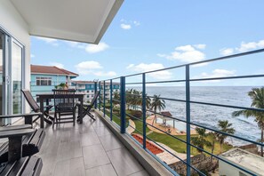 Un balcon avec vue sur la mer, une table à manger avec des chaises et un immeuble à la façade bleue.