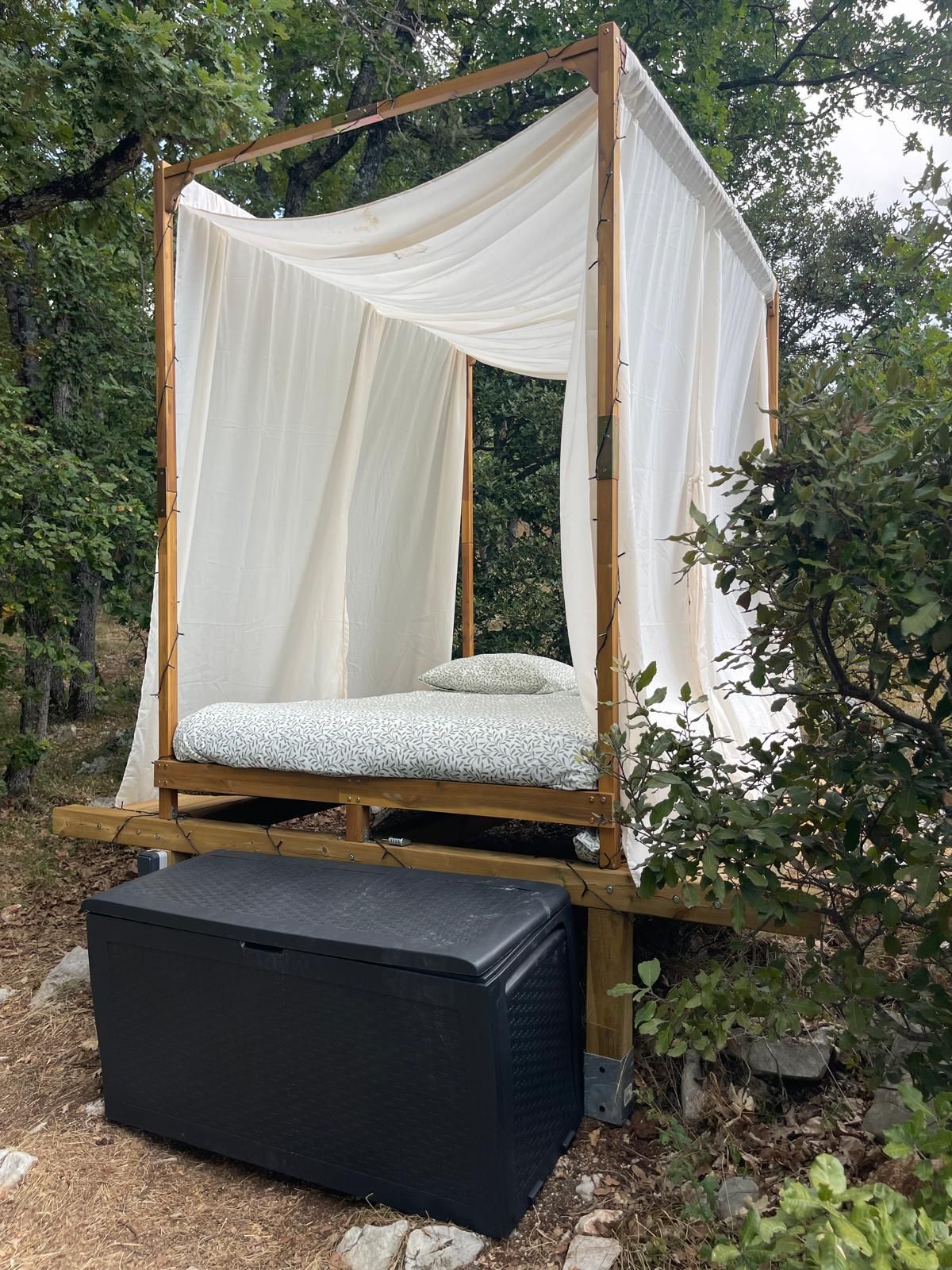 Een houten bed met hemel, een matras met een patroon en een zwarte opbergdoos.