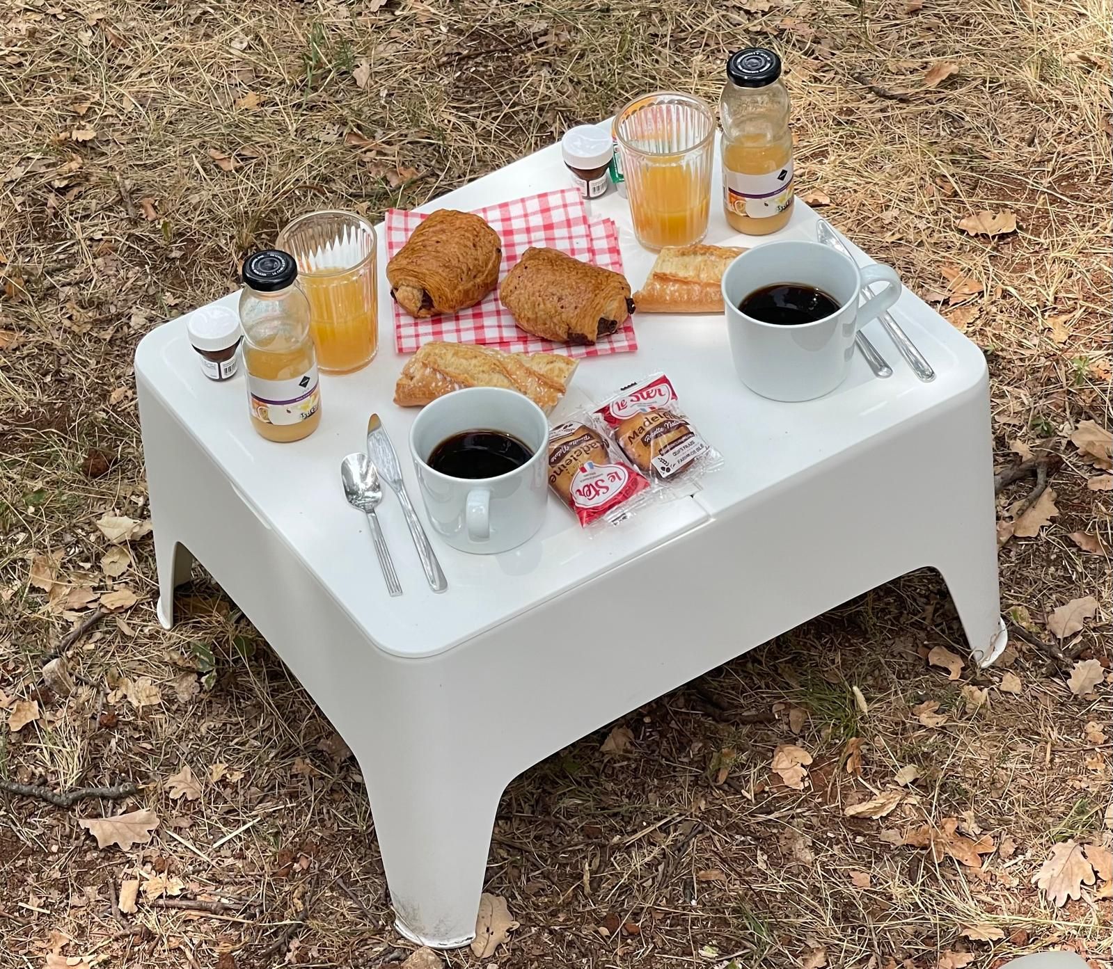 Een picknickopstelling met een witte tafel, twee koffiekopjes, een glas sap, croissants en snacks in verpakking, uitgestald op een geruit kleed in de open lucht.