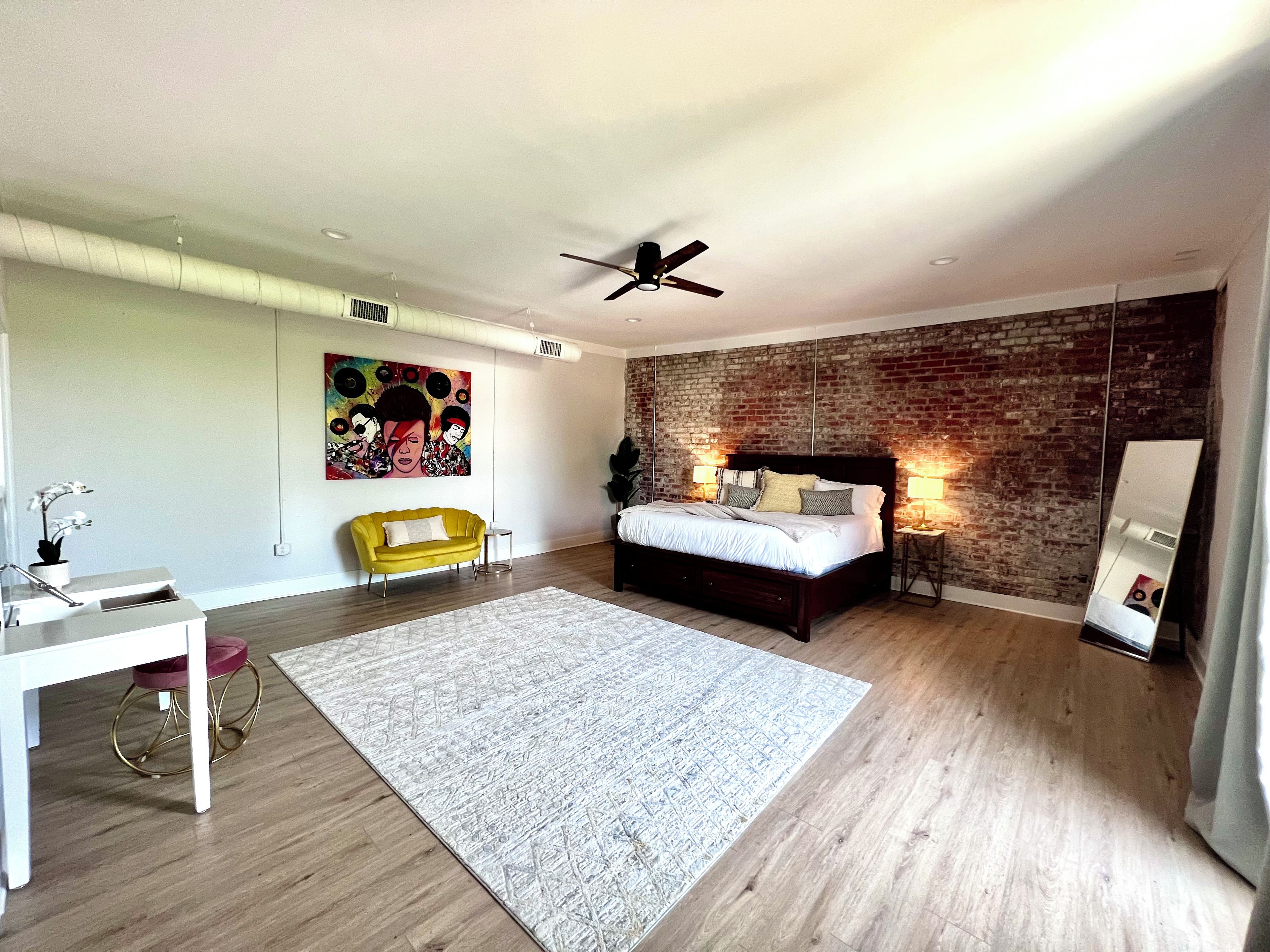 A modern bedroom with a brick wall, a bed with pillows, a yellow armchair, and a white desk.