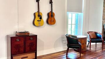 A room with wooden flooring, a dark wooden cabinet, two guitars on the wall, and a window with blinds.