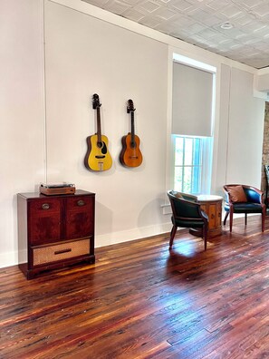 A room with wooden flooring, a dark wooden cabinet, two guitars on the wall, and a window with blinds.