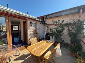 Une terrasse avec une table et des chaises en bois, un fauteuil en osier, des plantes en pot et un mur en pierre.