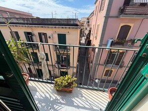 Une rue étroite en milieu urbain, bordée de bâtiments des deux côtés, un balcon orné d’un pot de fleurs et un ciel dégagé.