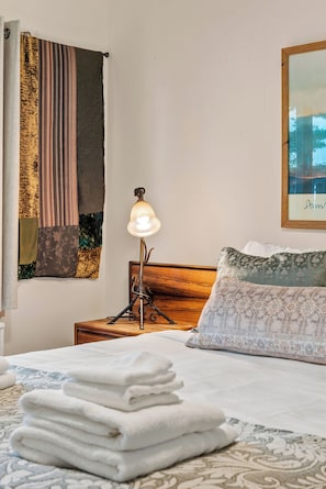 A neatly made bed with folded white towels, a bedside lamp, and a headboard with a floral pillow. - Private Beach | Game Room | Near Kohler-Andrae (Sheboygan)