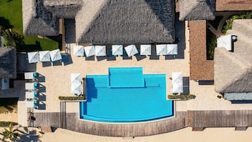 Eine Vogelperspektive zeigt ein Resort mit einem zentralen Schwimmbecken, strohgedeckten Gebäuden und Liegestühlen.