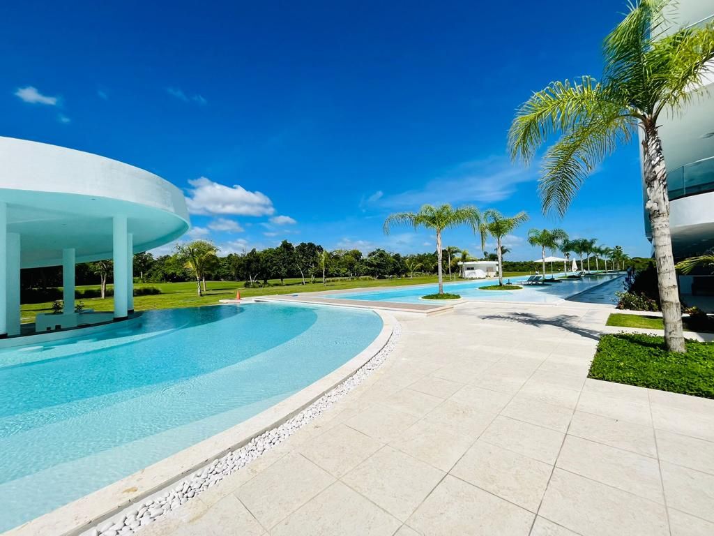 Un área de piscina moderna con una piscina de forma curva, un espacio de descanso cubierto y palmeras.