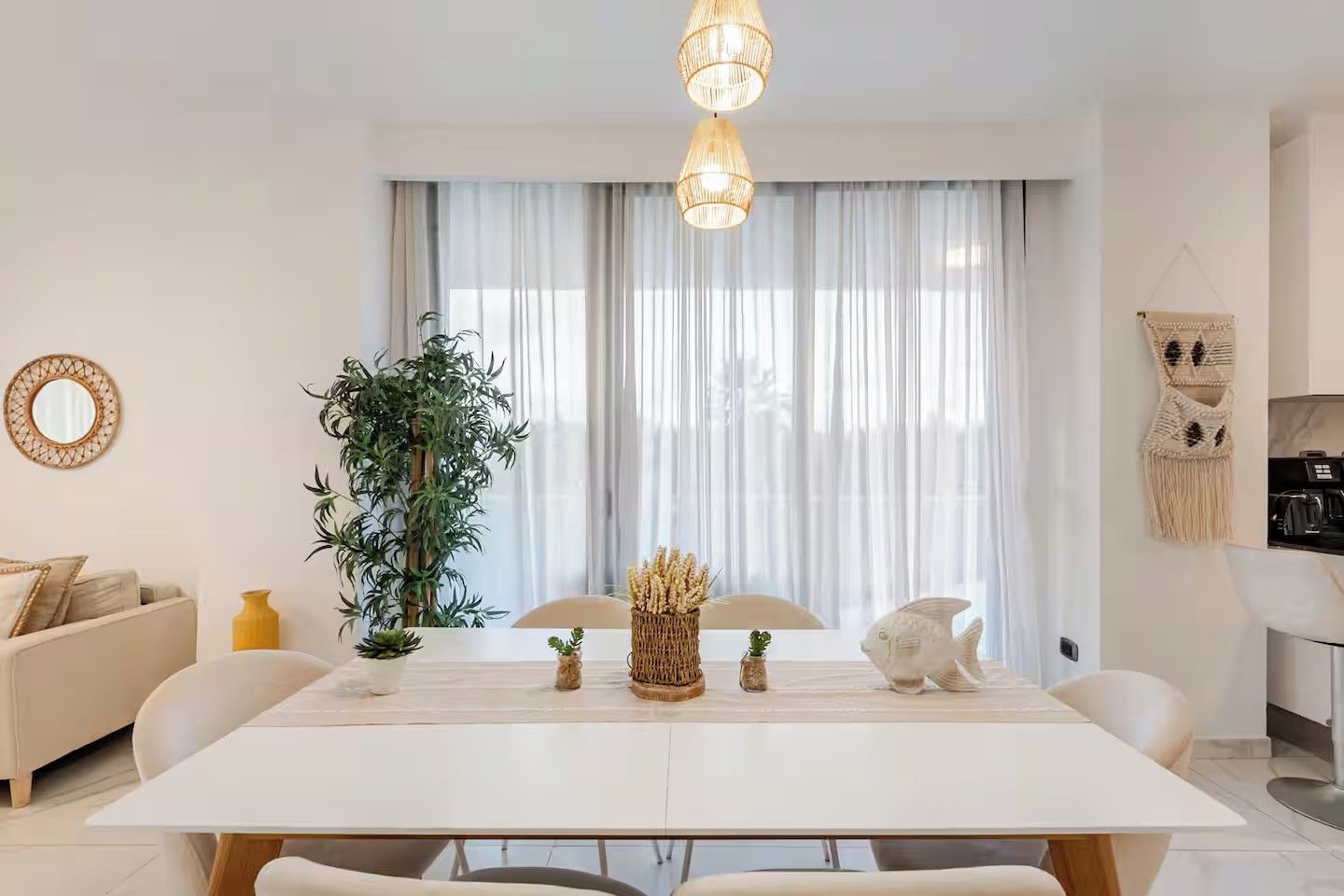 Ett modernt vardagsrum med ett vitt matbord, en soffa, ett stort fönster med tunna gardiner, en hängande lampa och ett köksområde som syns till höger.