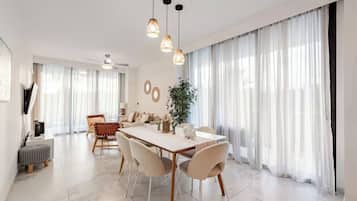 Un espace de salle à manger moderne avec une table blanche, des chaises beige et des luminaires suspendus.