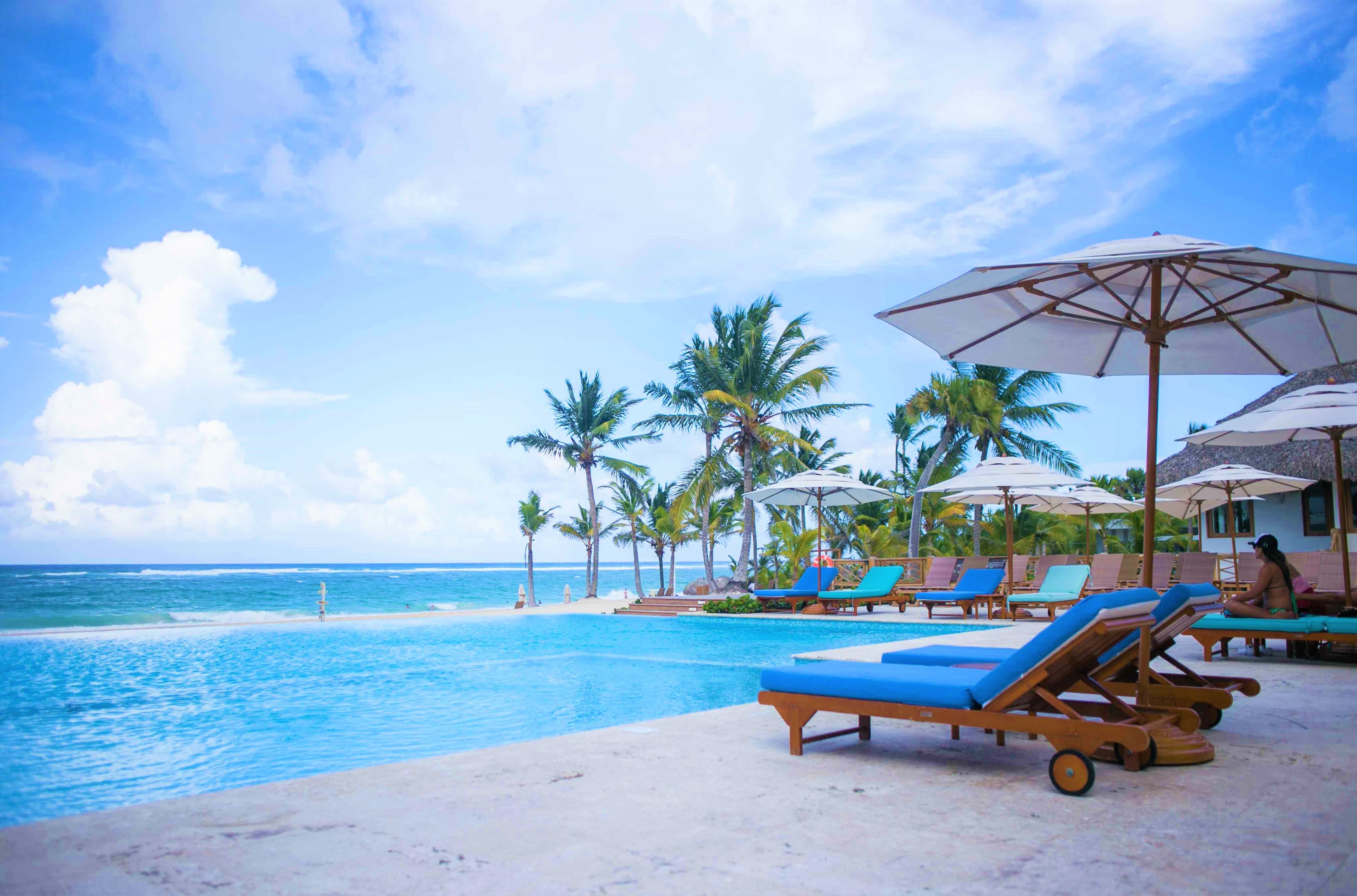 Un espace aménagé au bord de la piscine, avec des chaises longues et des parasols, des palmiers et une vue sur l’océan.