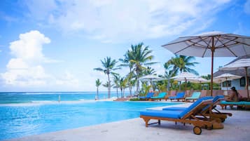 Un espace aménagé au bord de la piscine, avec des chaises longues et des parasols, des palmiers et une vue sur l’océan.