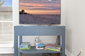 Un televisor de pared mostrando un atardecer en la playa, un aparador azul con juegos de mesa y un enchufe.