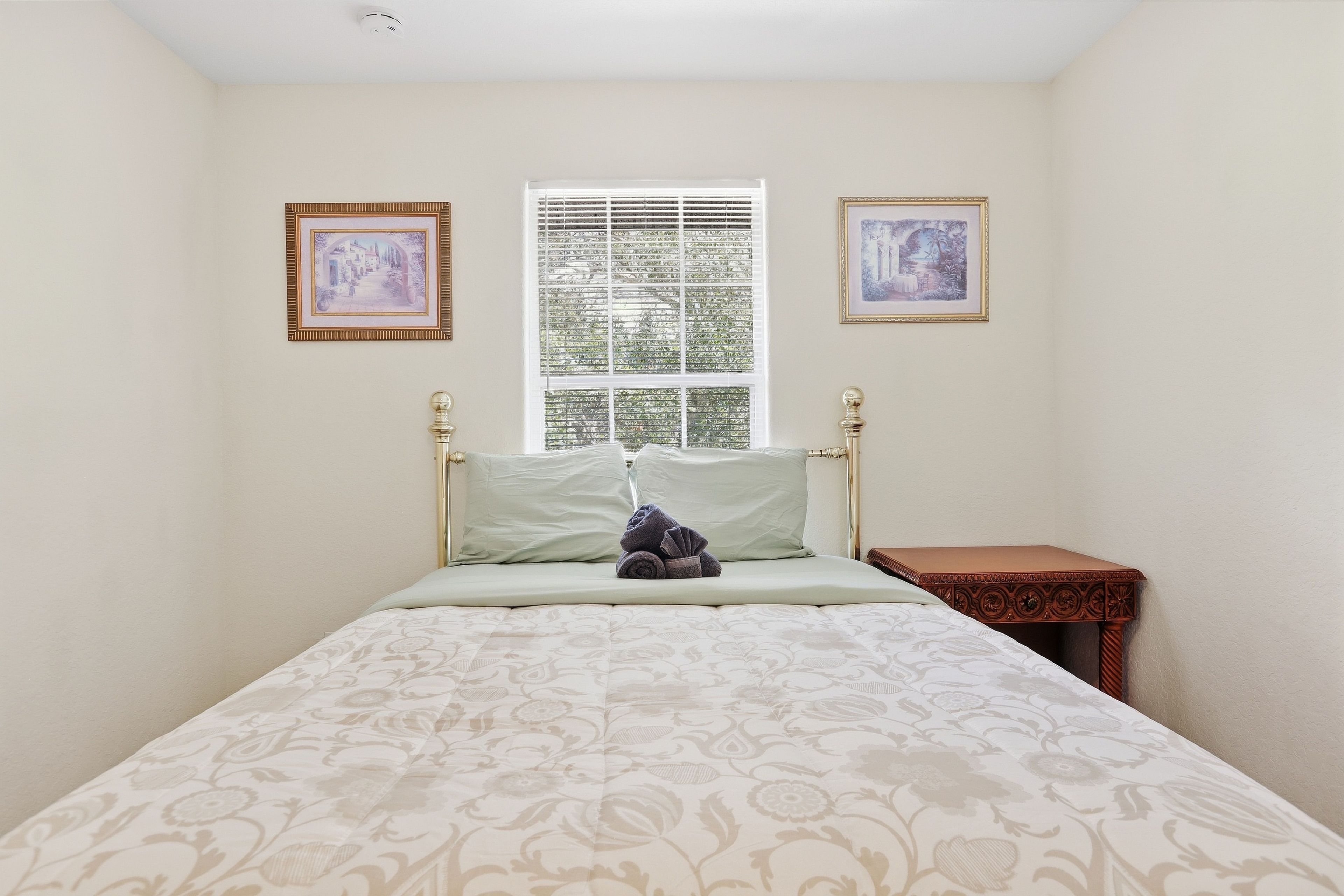 A bedroom with a bed, a window with blinds, two framed pictures on the wall, and a small wooden nightstand.