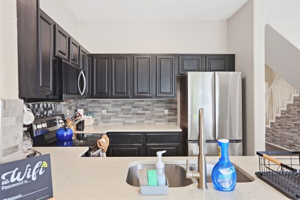 A modern kitchen with dark cabinets, a stainless steel refrigerator, and a countertop with a sink and various kitchen items.