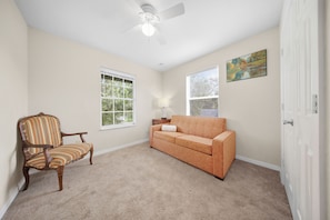 A room with a ceiling fan, a window with blinds, a sofa, a chair, and a painting on the wall.