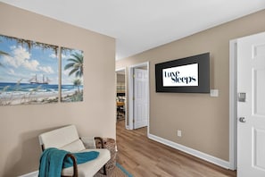 A hallway with a beige wall, a framed painting of a beach scene, a sign reading 'Luxe Sleeps', and a door leading to a room. - Savannah Reserve Savannah Breeze Suite & Hot Tub (Savannah)