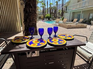 A poolside area with lounge chairs, a table with decorative plates and blue glasses, and a view of a pool and palm trees. - Downtown Mid-Century Gem with Pool & Mountain Views, Heated Spa, Sleeps 4! (Palm Springs)