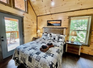 A wooden cabin bedroom with a bed, two nightstands, a painting, and a door leading to an outdoor deck. - Bearly Behavin | Hot Tub Pool Table Great Location (Sevierville)