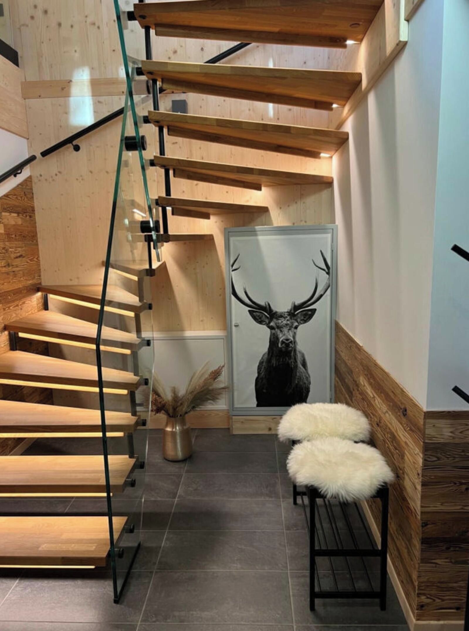Un escalier moderne avec des marches en bois et une rampe en verre, menant à une pièce décorée d’une affiche représentant un cerf et d’un vase contenant des plantes séchées.