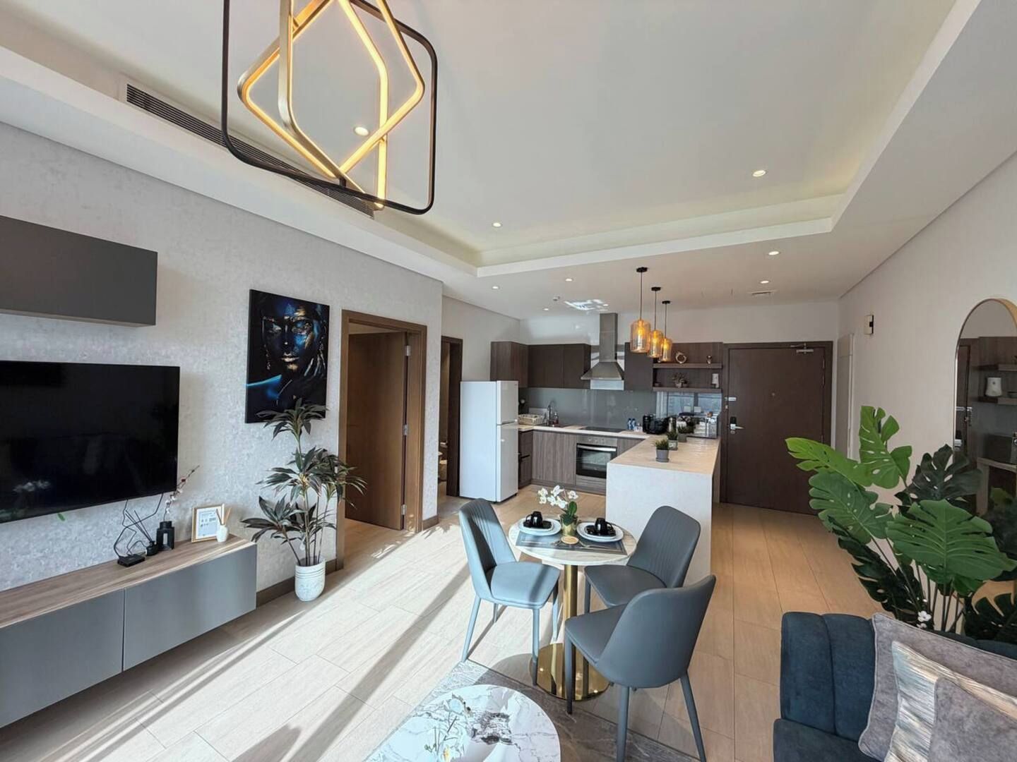 Uma sala de estar moderna com televisão de tela plana, área de jantar com mesa redonda e cadeiras, cozinha com ilha central e uma grande planta em vaso.