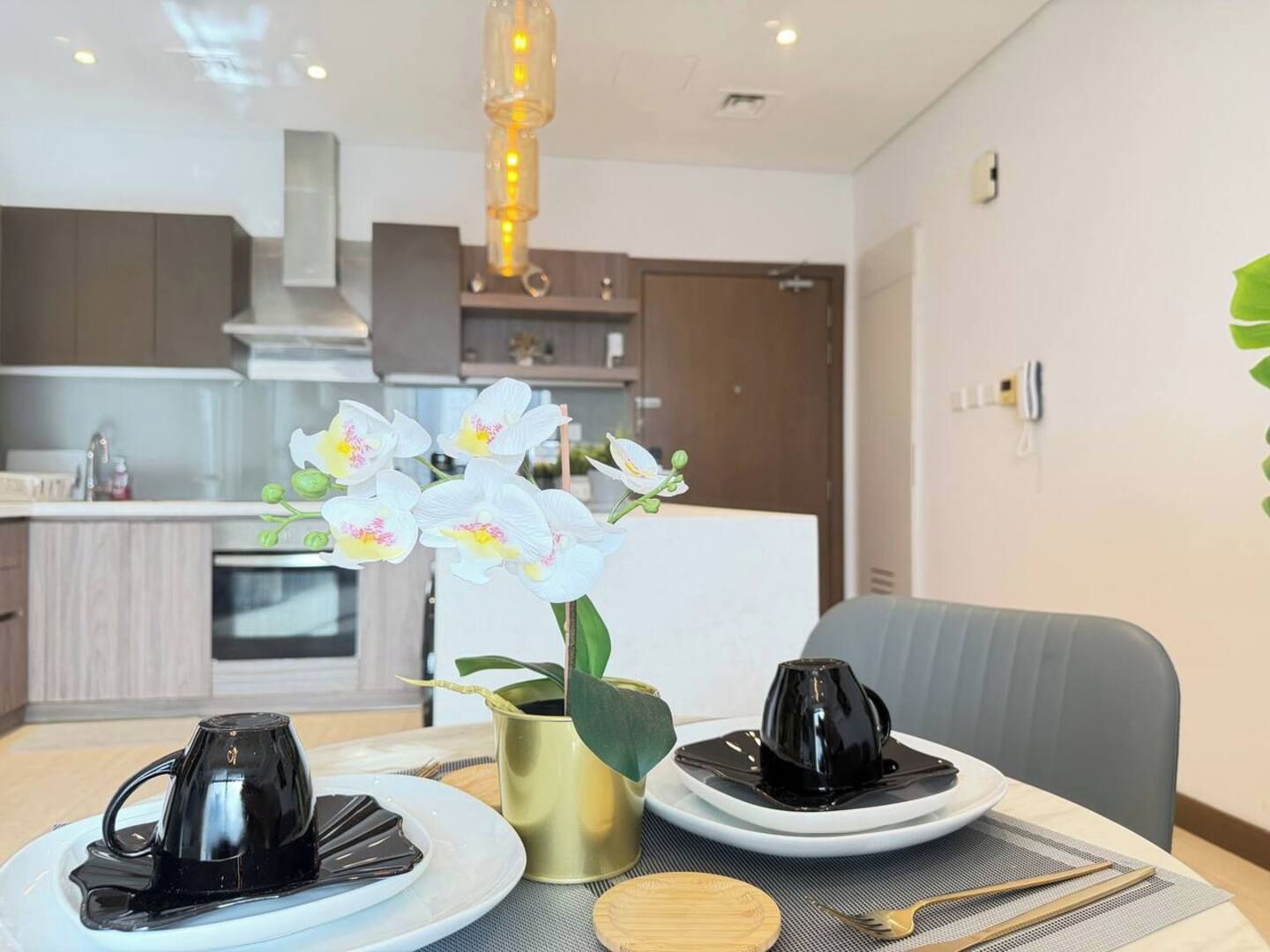 Un coin repas moderne avec une table dressée pour deux personnes, agrémenté d’une cafetière, d’un vase avec des fleurs blanches et d’une pièce de pâtisserie noire présentée sur des assiettes.