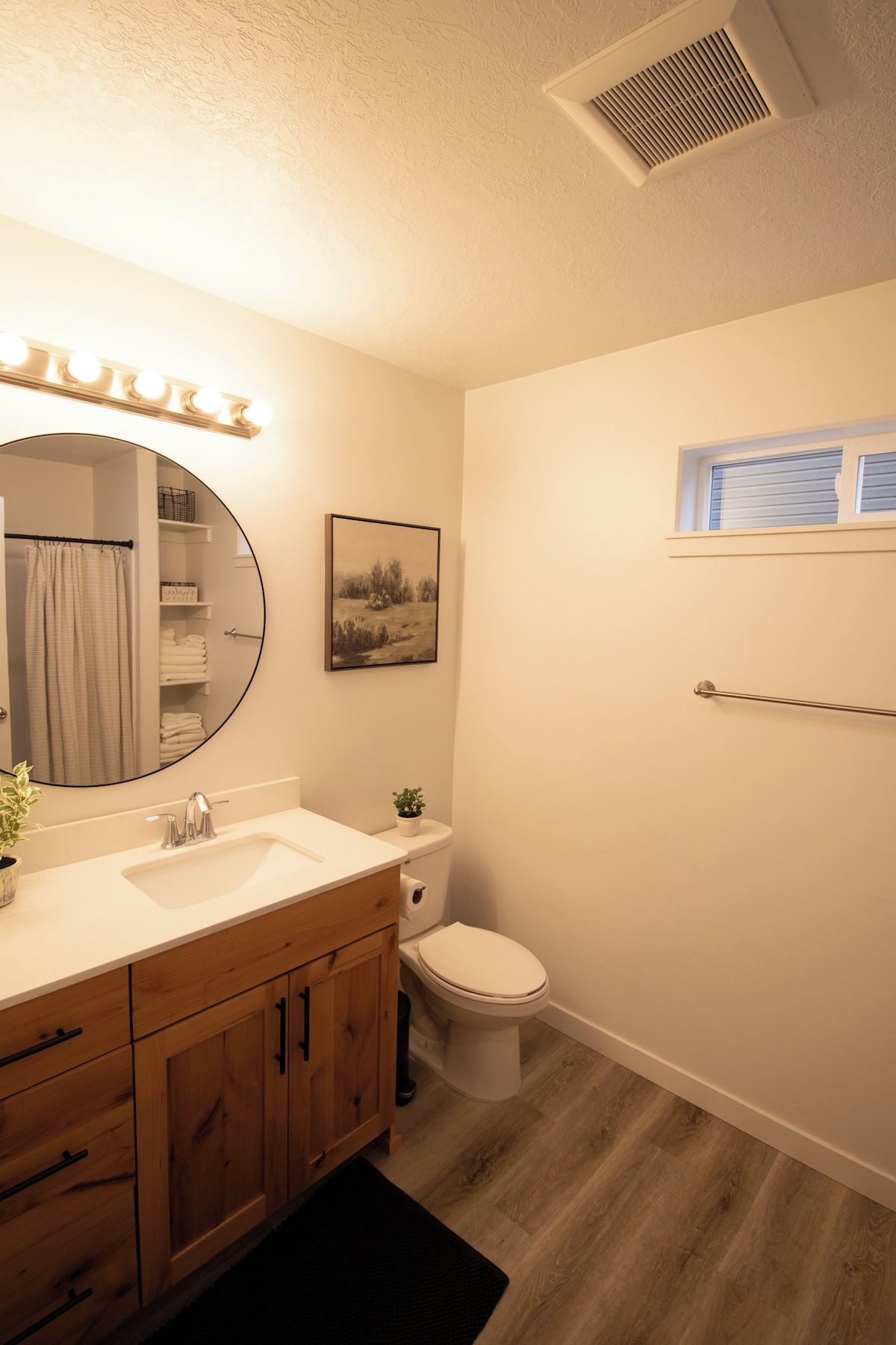Une salle de bain comprenant un meuble-lavabo, des toilettes, un rideau de douche, un porte-serviettes, une fenêtre et un tableau accroché au mur.