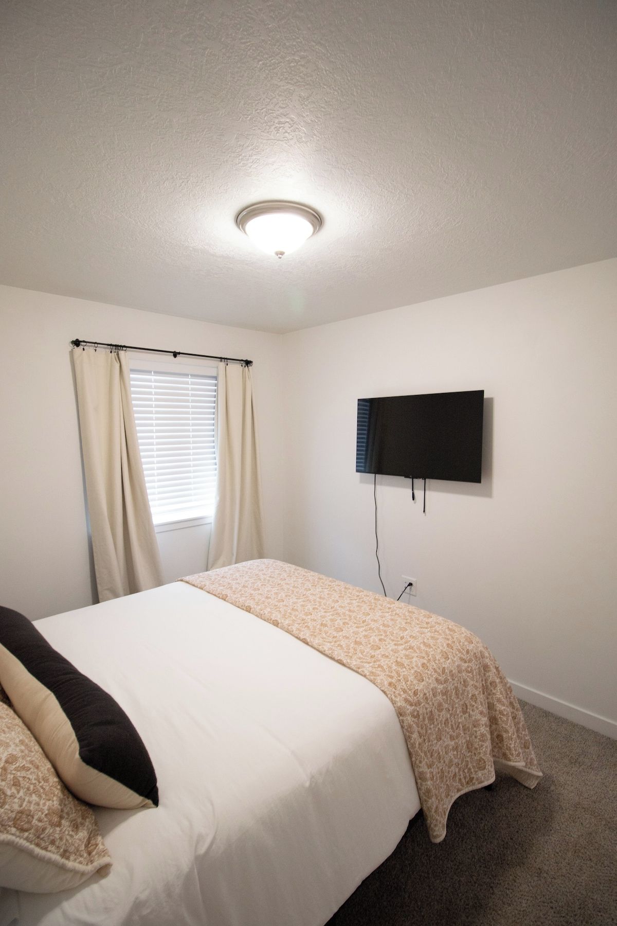 A bedroom with a bed, curtains, a television mounted on the wall, and a ceiling light.