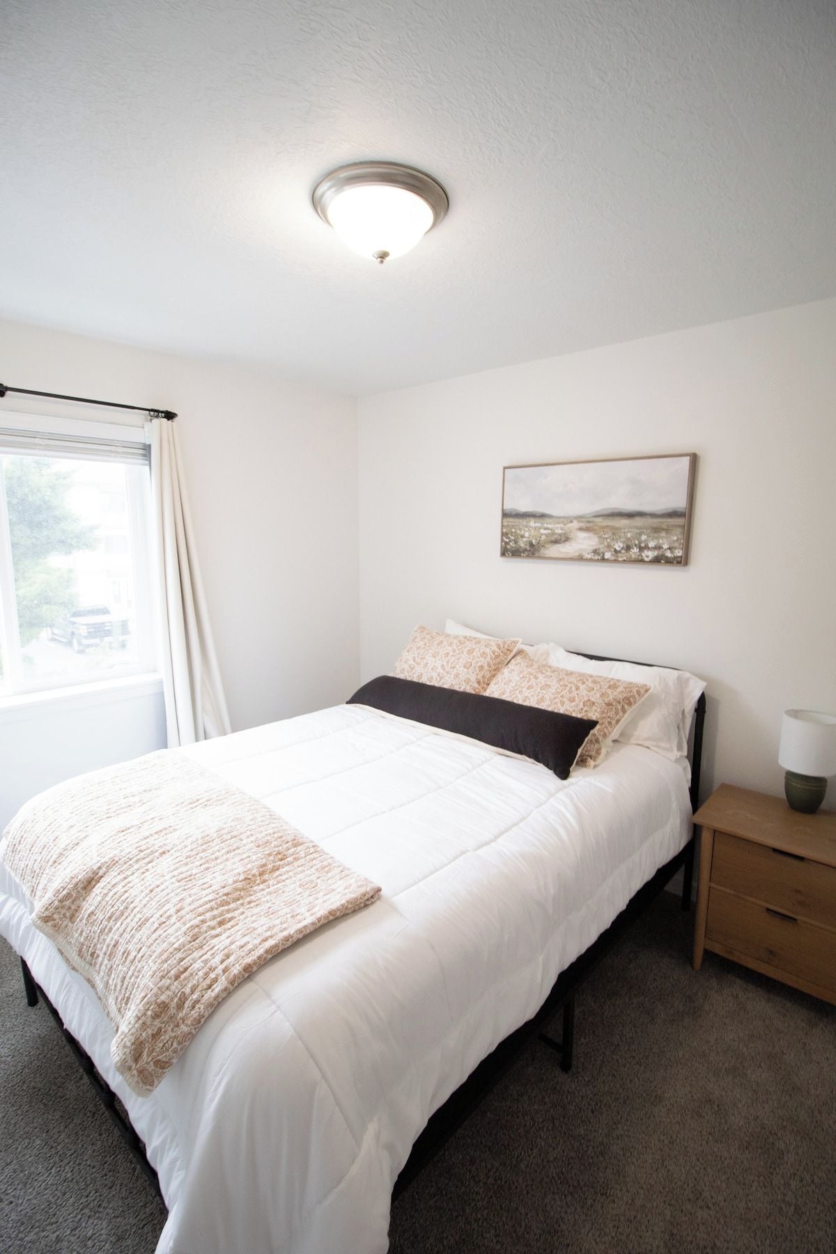 A neatly made bed with a white duvet and a brown throw blanket, a bedside table with a lamp, and a window with curtains.