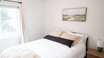 A neatly made bed with a white duvet and a brown throw blanket, a bedside table with a lamp, and a window with curtains.