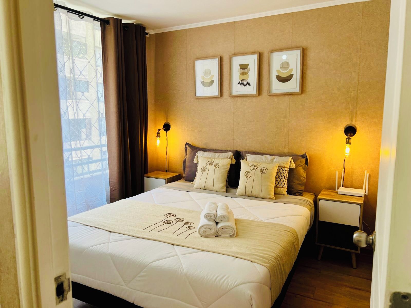 Une chambre d’hôtel avec un grand lit, deux tables de chevet, une lampe fixée au mur et une fenêtre avec des rideaux.