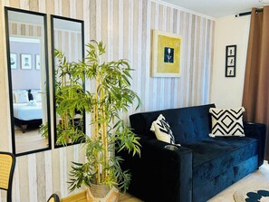 A living room with a dark blue sofa, a large potted plant, and a mirror reflecting a bedroom. - El mejor lugar - Hospital UC - Metro (Santiago)