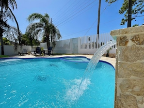 A swimming pool with a waterfall feature, surrounded by a white fence, palm trees, and lounge chairs. - Charming Beach Home Dorado Del Mar and Golf (Dorado)