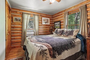 Un dormitorio de cabaña de troncos con una cama, paredes de madera, una ventana y un ventilador de techo.