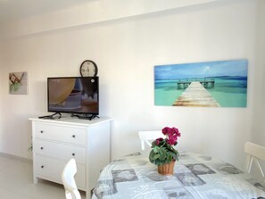 A room with a television on a white cabinet, a painting of a beach, a wooden table with a potted plant, and a white chair. - Apartment in Nerja Near Balcon de Europa (Nerja)