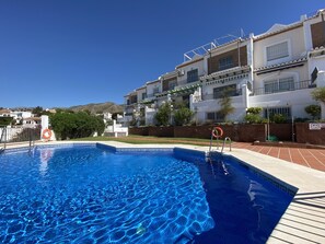 Ein Swimmingpool mit glatter, klarer blauer Wasseroberfläche, umgeben von einem weißen Gebäude mit Balkonen und einem grünen Zaun.