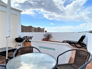 Eine Dachterrasse mit Glastisch, Korbstühlen, einem Grill, Topfpflanzen und Meerblick.