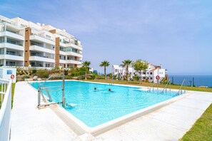 Ein Swimmingpool mit klarem, blauem Wasser, umgeben von einem weißen Gebäude mit mehreren Balkonen und Blick aufs Meer.