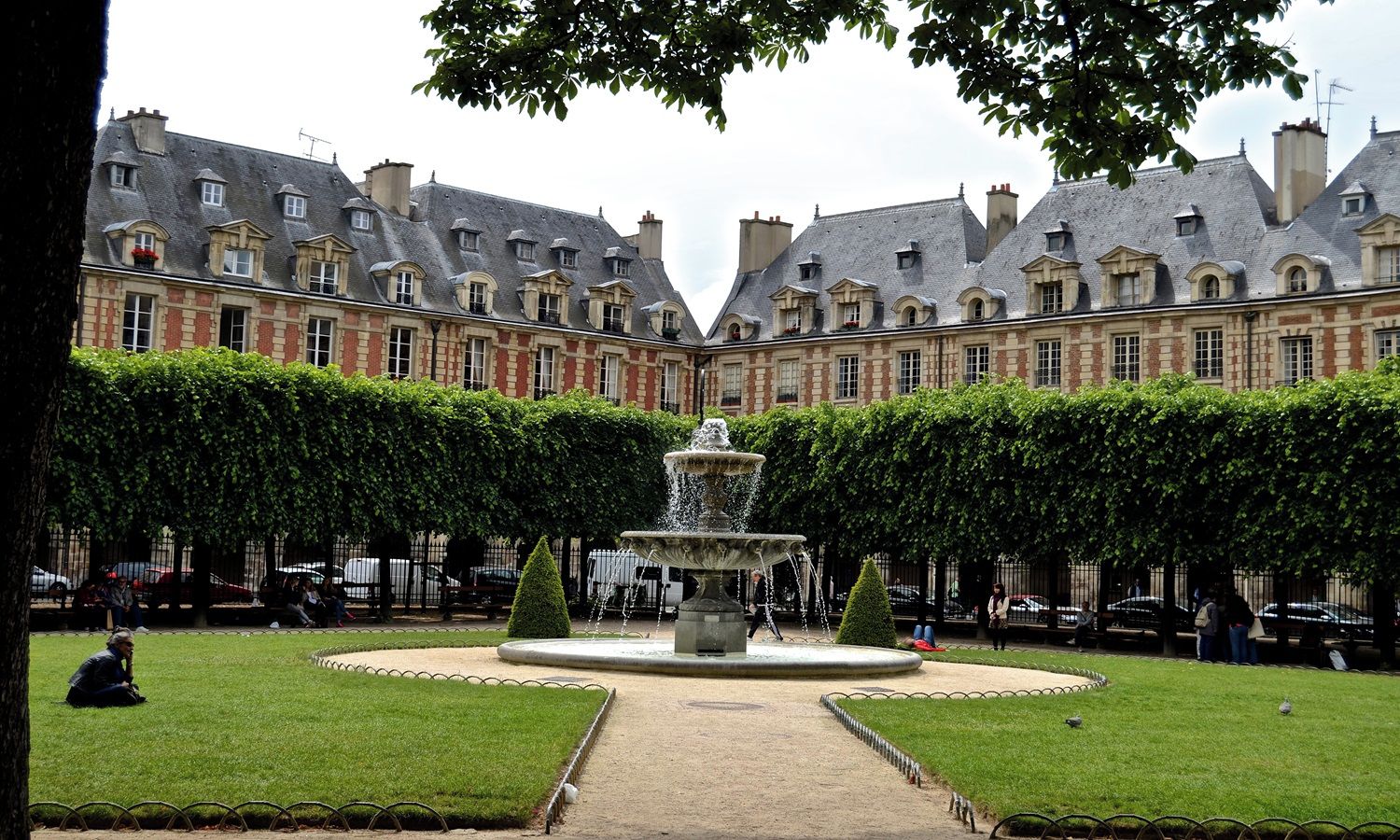 A courtyard with a fountain, surrounded by a hedge and a building with multiple windows and chimneys.