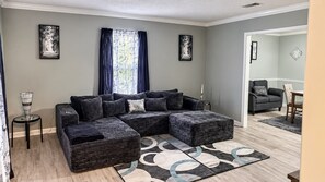 Ein Wohnzimmer mit einem Ecksofa, einem Couchtisch und einem Teppich. Es gibt ein Fenster mit Vorhängen, einen Beistelltisch und eine Tür zu einem anderen Raum.