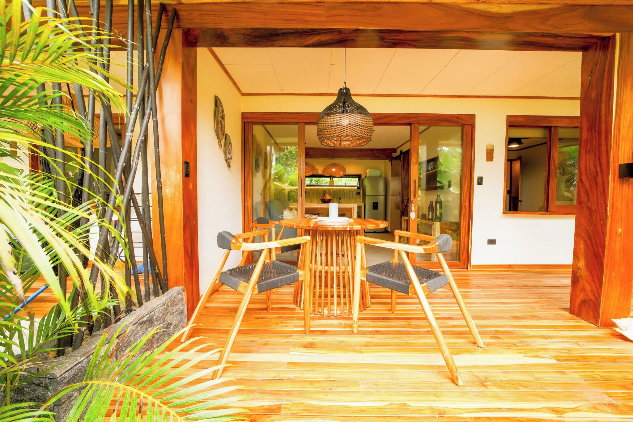 Quarto com piso de madeira, uma mesa redonda e cadeiras, luminária pendente e vista para outro cômodo com uma geladeira.