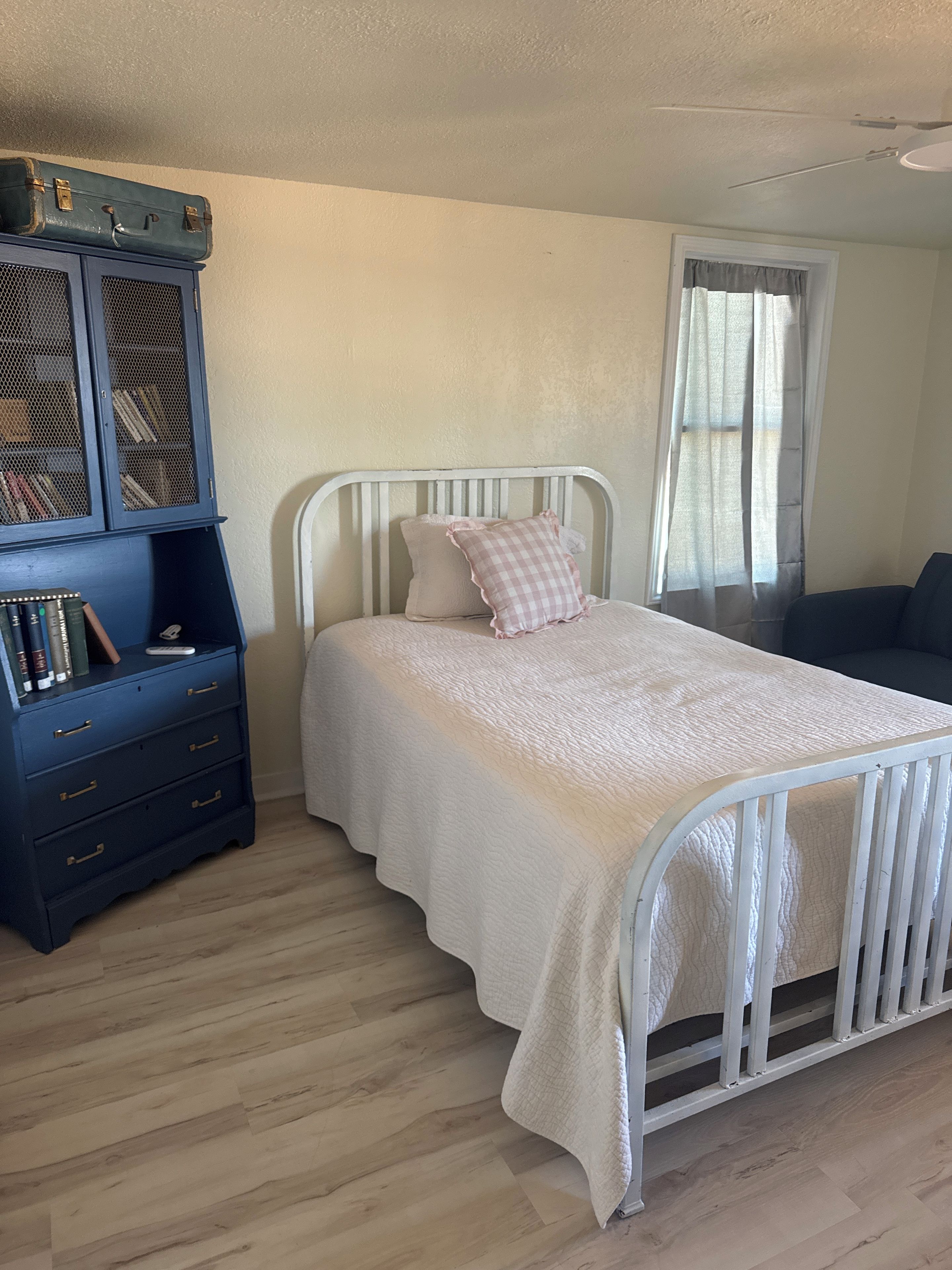 A bedroom with a white bed, a blue dresser with books, and a window with curtains.