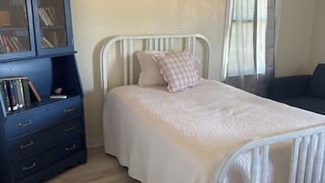 A bedroom with a white bed, a blue dresser with books, and a window with curtains.