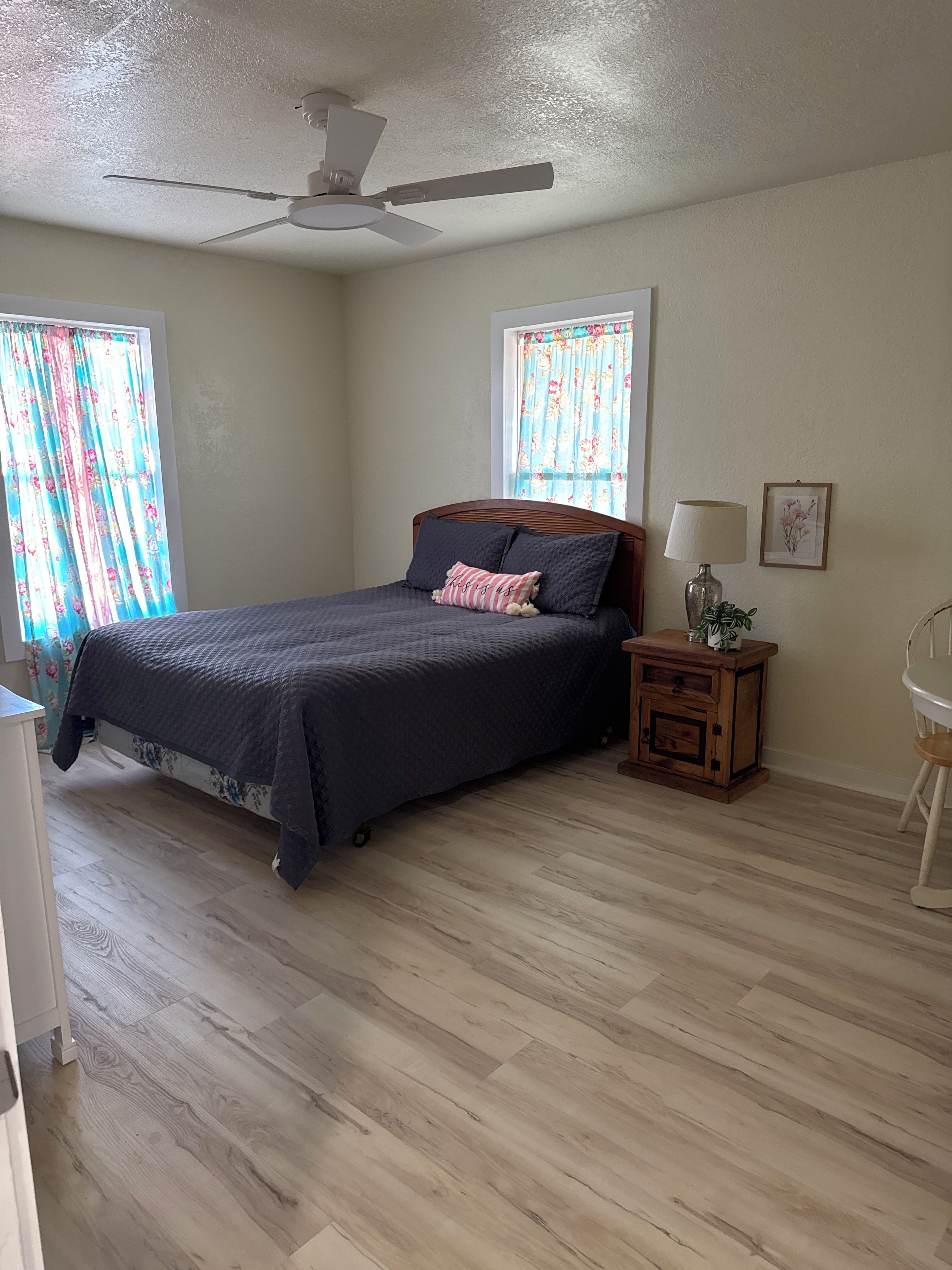 A bedroom with a bed, a ceiling fan, a nightstand with a lamp, and a window with curtains.
