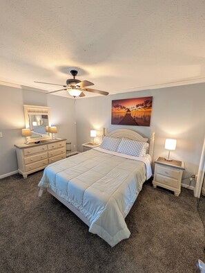 Een slaapkamer met een bed, nachtkastjes, een dressoir, een plafondventilator en een schilderij van een zonsondergang aan de muur.