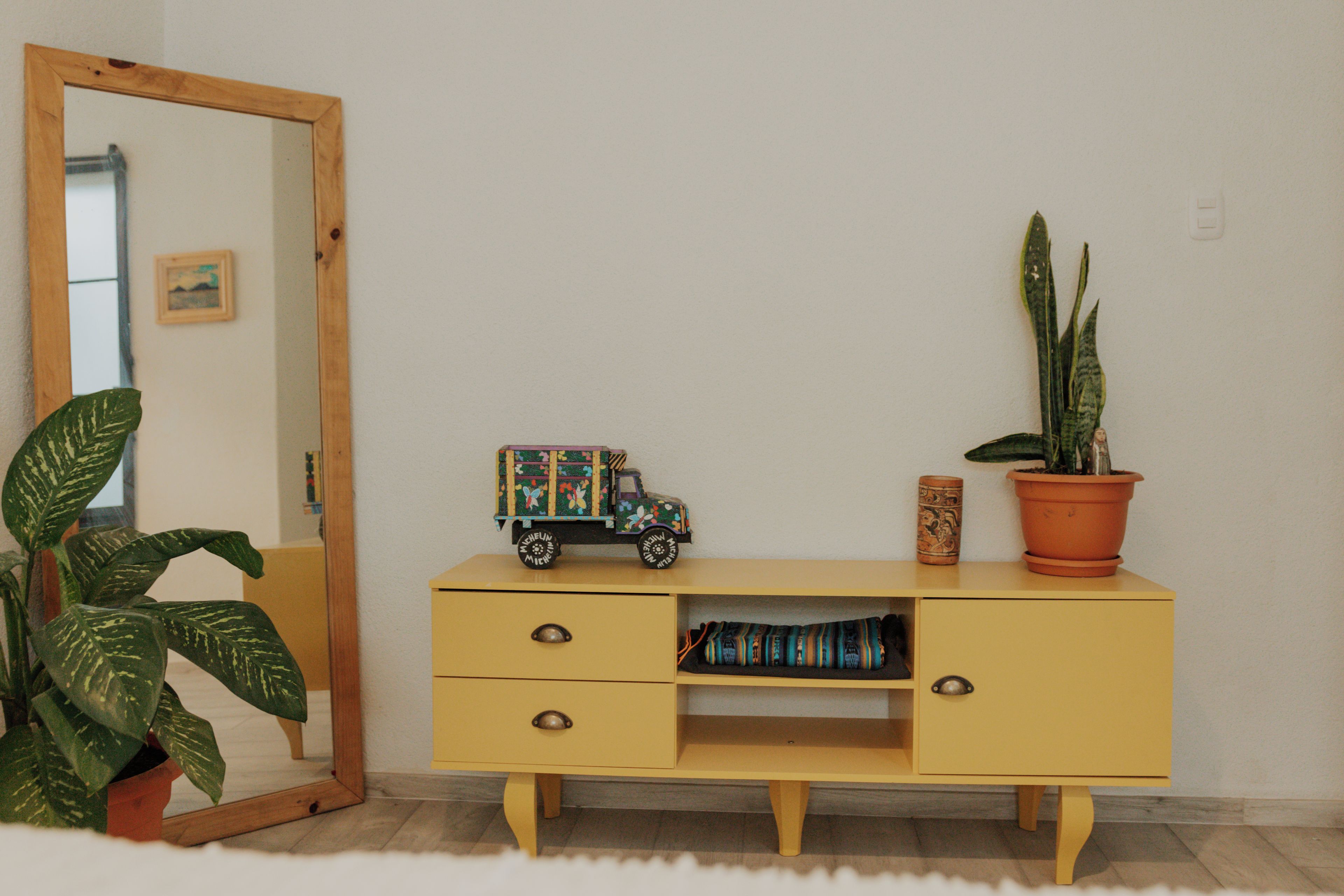 Una habitación con un aparador amarillo, un espejo, un camión de juguete, una planta en maceta y un pequeño objeto decorativo.