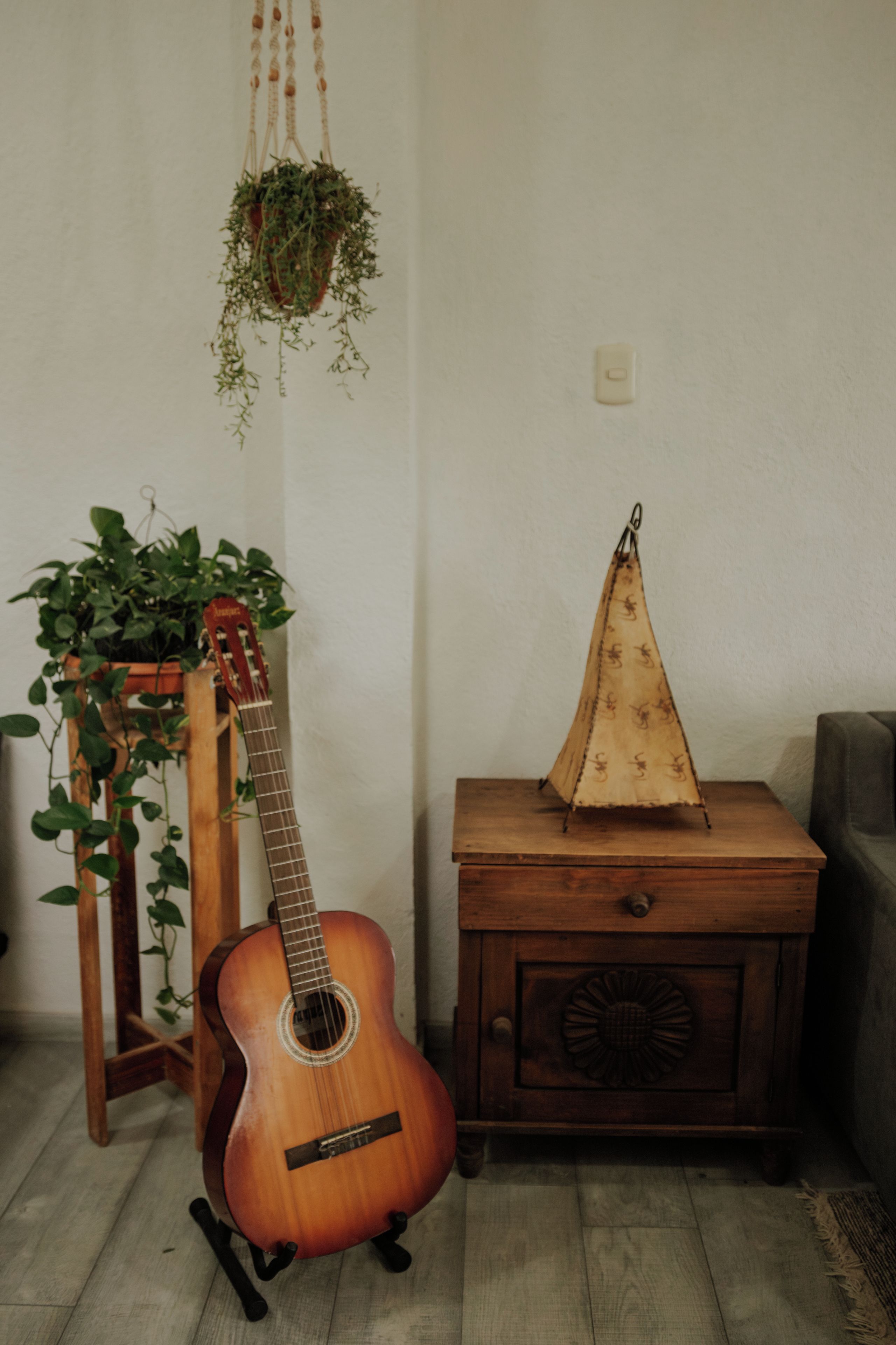 A guitar on a stand, a wooden nightstand with a decorative item, and a hanging plant.